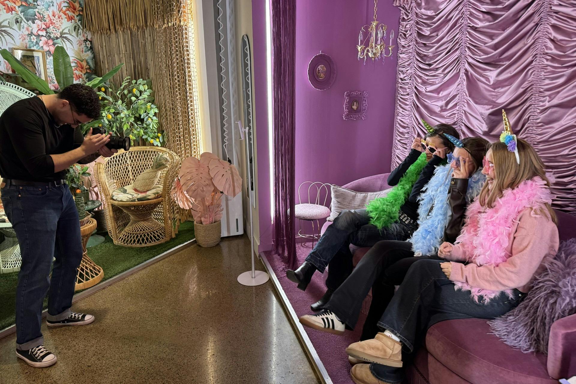 Τρία άτομα ντυμένα με πολύχρωμα κασκόλ και κορδέλες μονόκερου κάθονται σε έναν μοβ καναπέ σε ένα δωμάτιο με ζωηρή μοβ διακόσμηση.