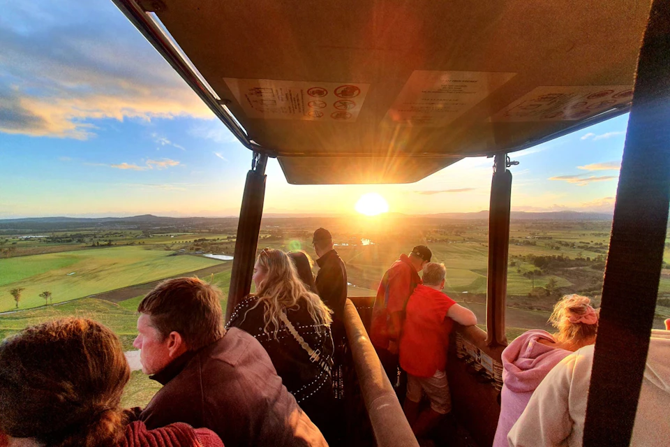 People in a hot air balloon basket enjoy views of a vast green landscape during sunset.