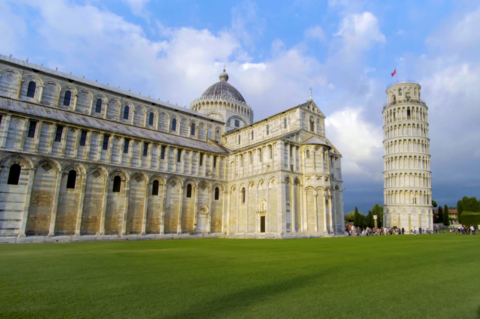A historic cathedral with the Leaning Tower of Pisa in the background under a partly cloudy sky, with a green lawn in the foreground.
