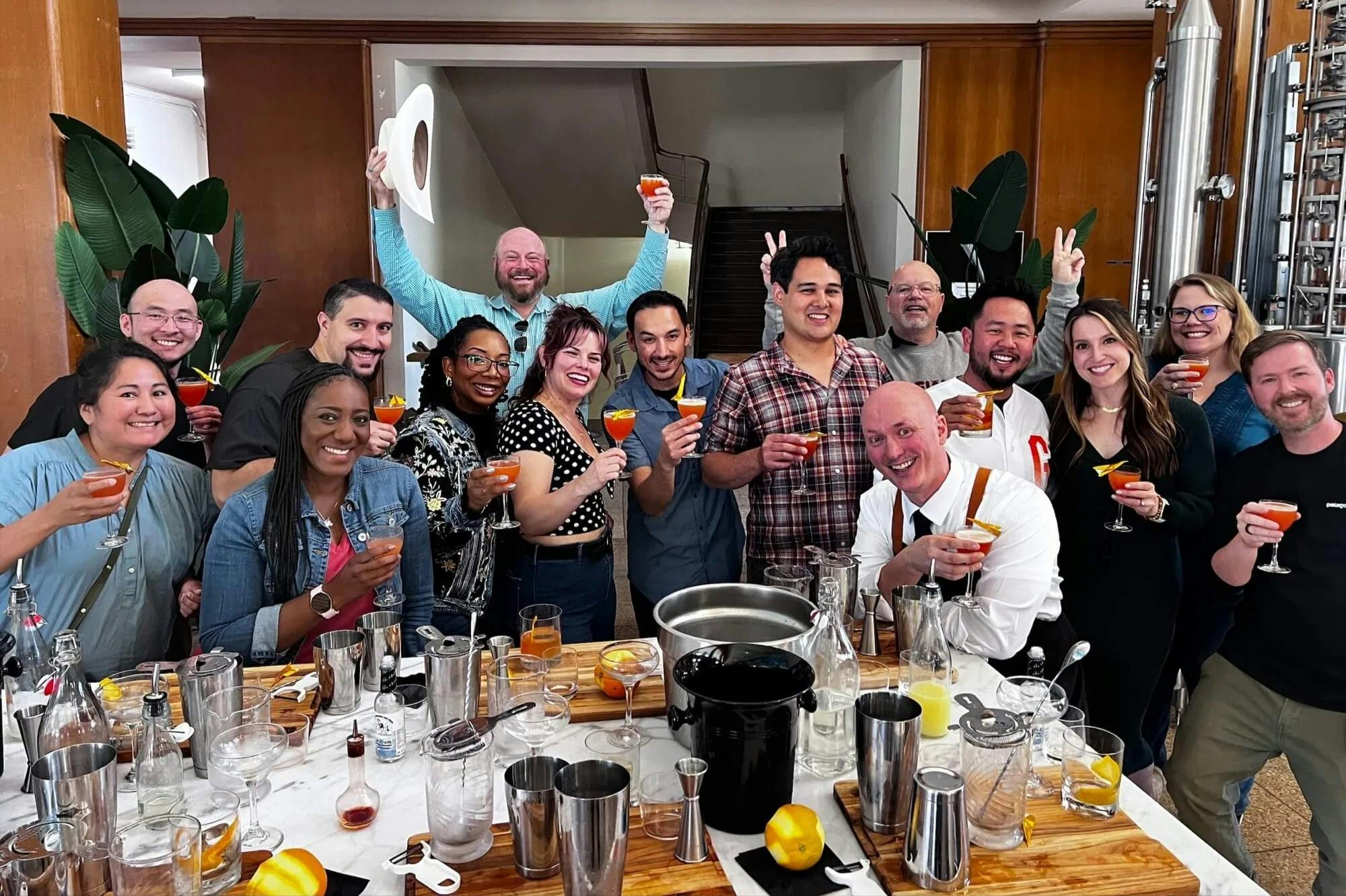 A group of people standing around a wooden table holding cocktails, smiling, with bartending tools and glasses on the table.
