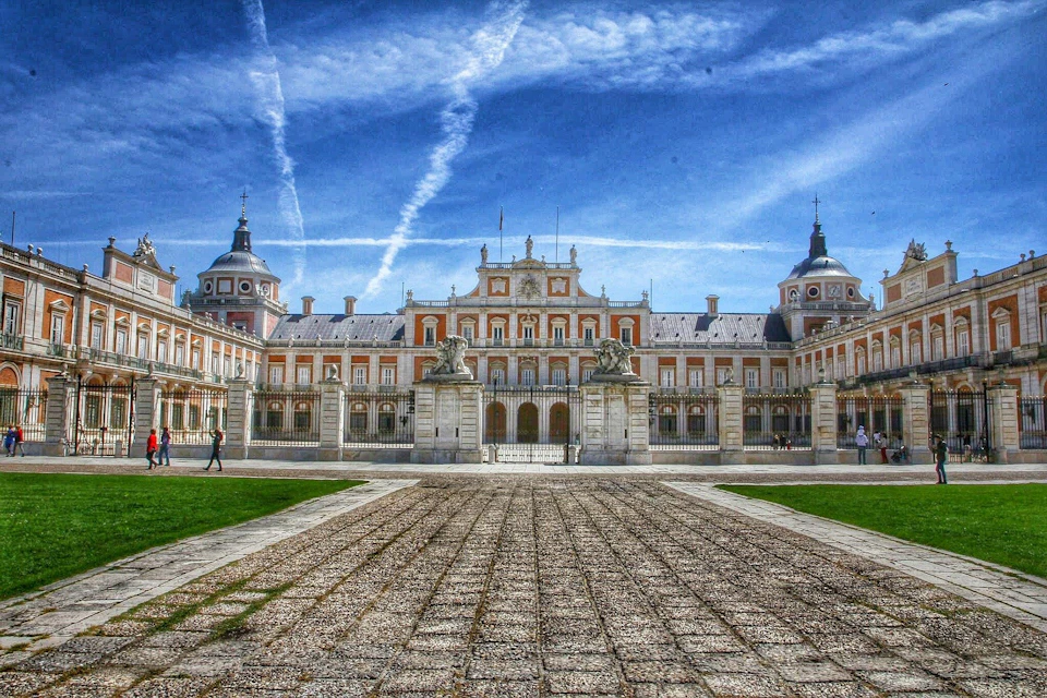 Ornate palace with symmetrical architecture, a central entrance flanked by statues, under a blue sky with contrails.