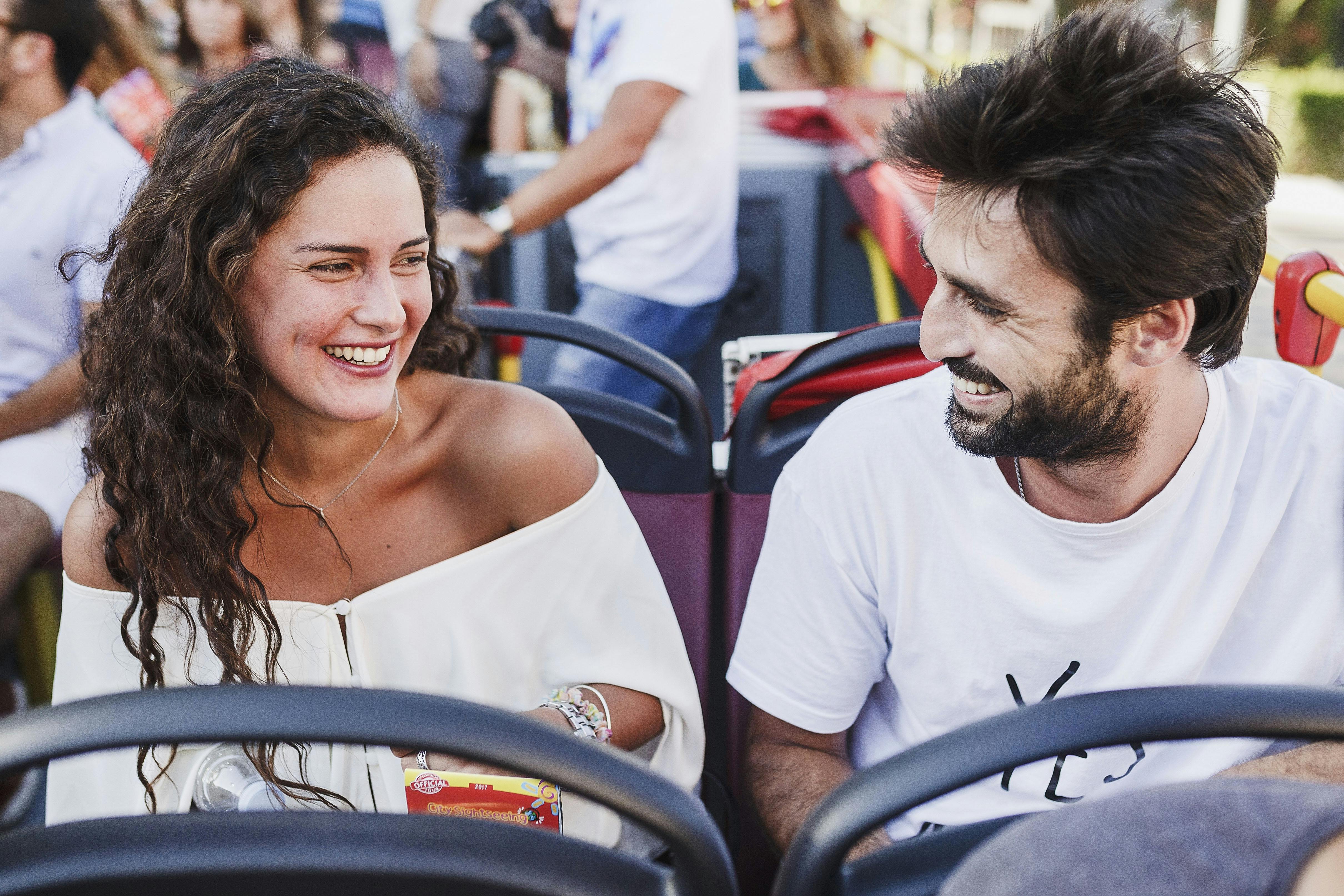 Dva lidé se na sebe usmívají, zatímco sedí ve venkovním turistickém autobuse. Do jedné se vejde krabička s občerstvením. V pozadí jsou další cestující.