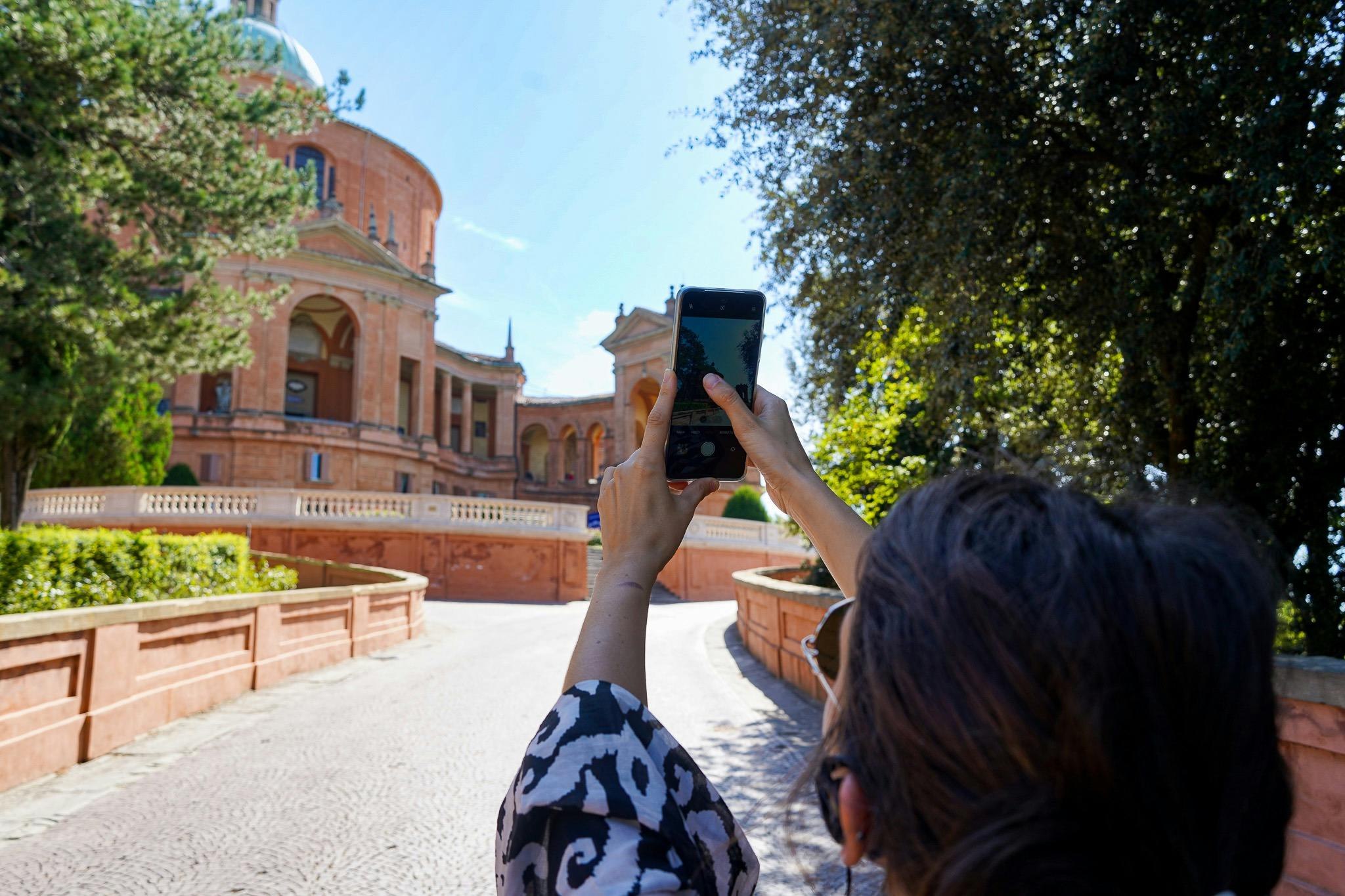 Person taking a photo of a historic, domed building with a phone. The scene is set on a sunny day with trees and a curved pathway.