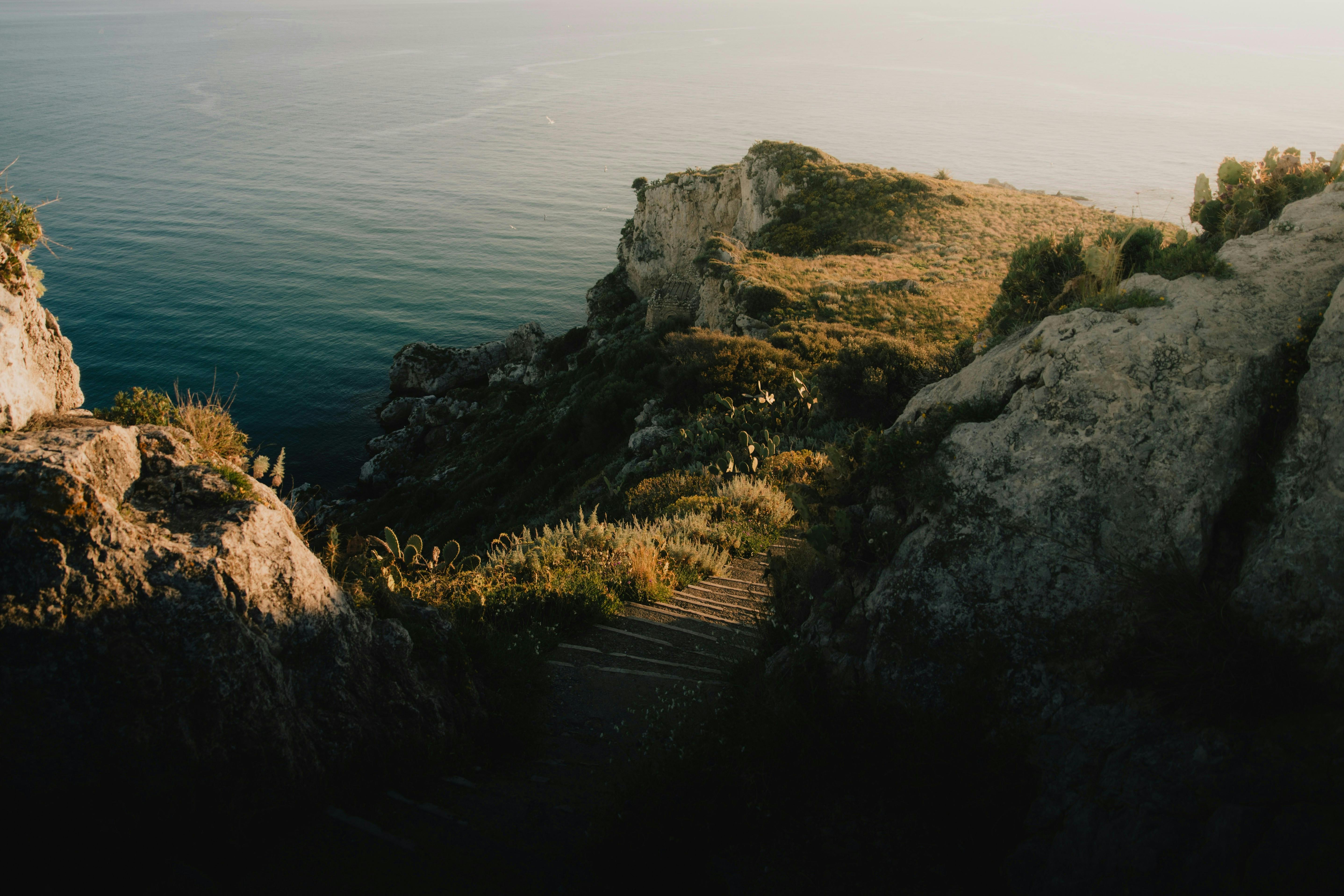 Stone stairs descend through rocky terrain with sparse vegetation, leading to calm ocean waters under a soft, golden sunset.