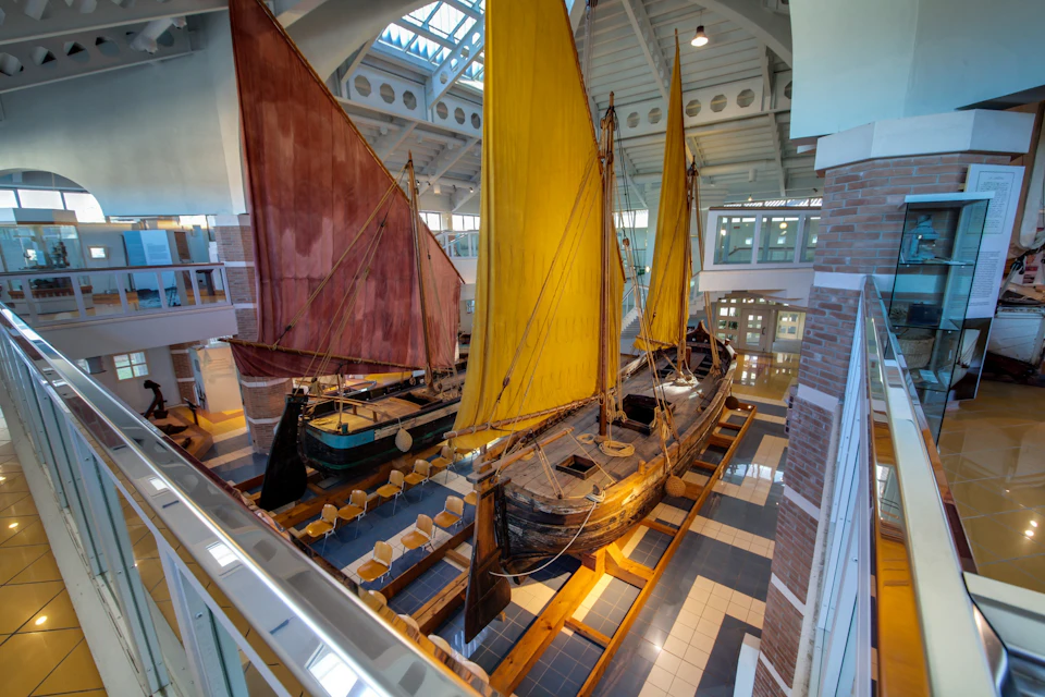 Two vintage sailboats with large red and yellow sails are displayed indoors on a tiled floor in a brightly-lit atrium.