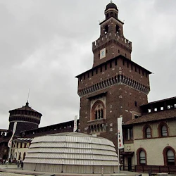 Historiskt tegeltorn med utsmyckade detaljer, omgivet av en innergård, modern vit struktur, människor och intilliggande byggnader under molnig himmel.