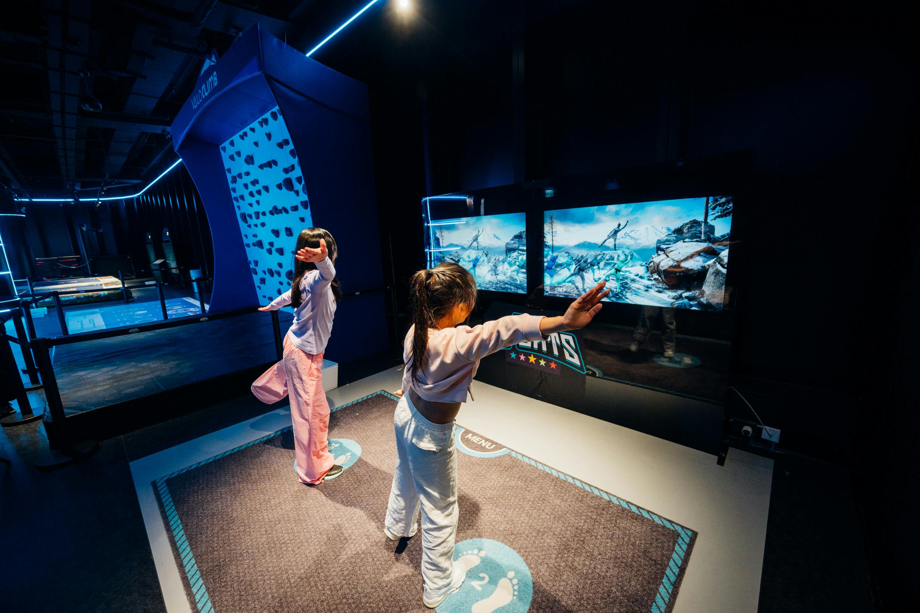 Two children stand on a gaming mat interacting with a virtual reality game displayed on two large screens in a dark room.