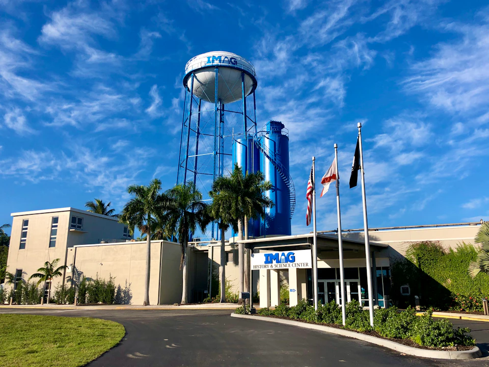 The entrance to IMAG History & Science Center with flags, palm trees, a blue water tower, and a clear sky.