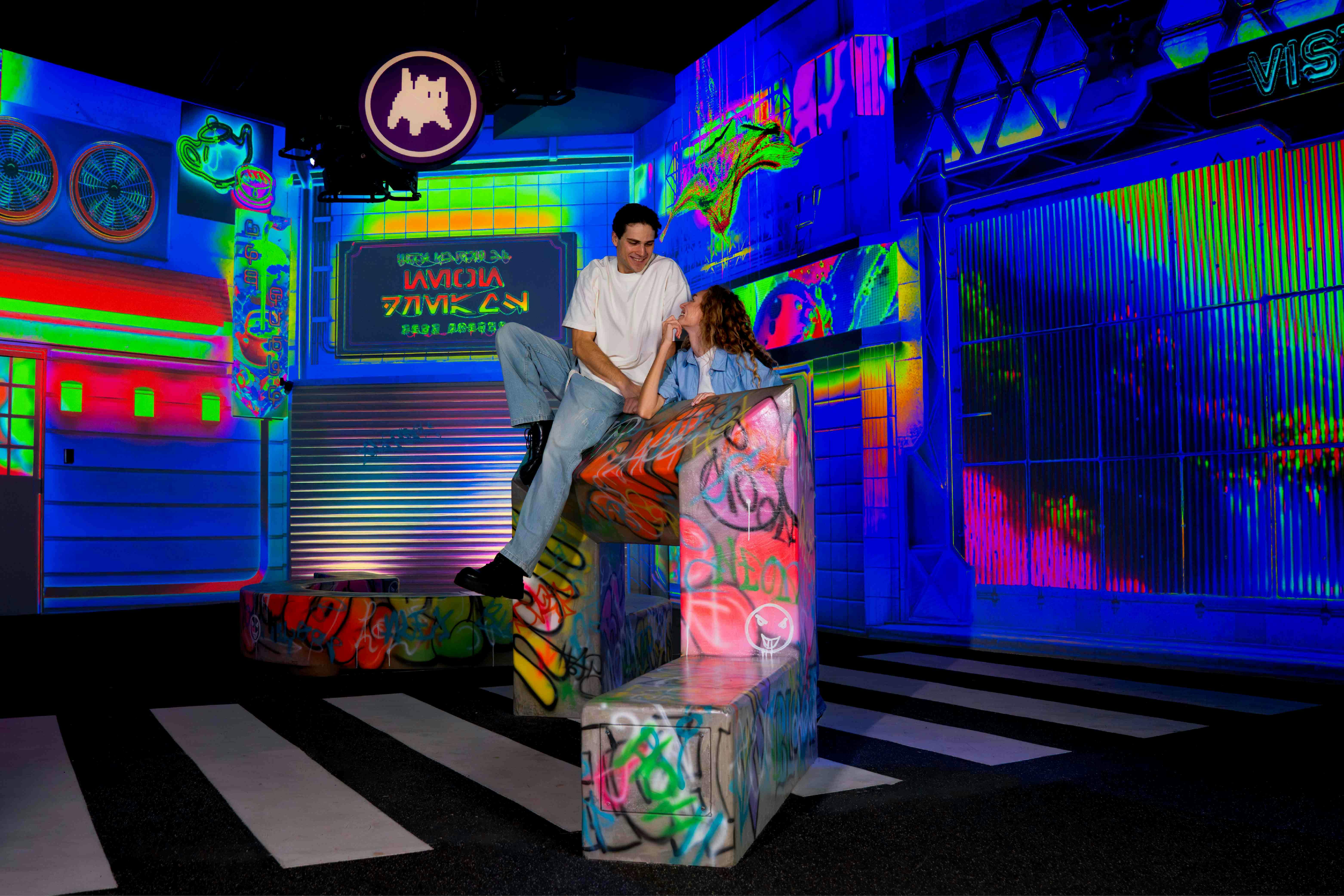 Two people sitting on graffiti-decorated structures in a neon-lit urban-themed environment.