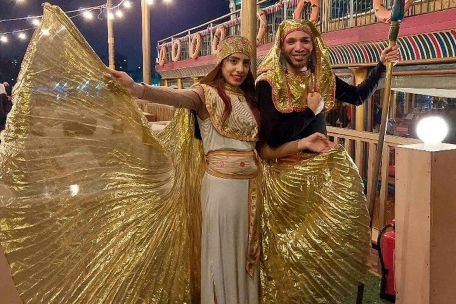 Dva lidé ve zlatých a bílých kostýmech v egyptském stylu pózují na palubě lodi se záchrannými kruhy a lanovými světly.