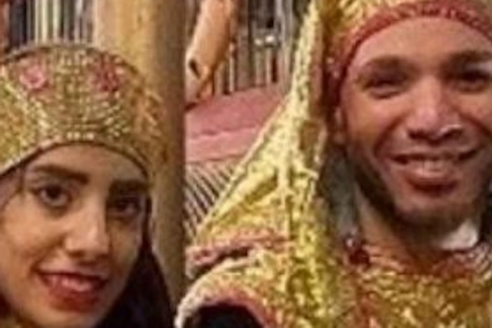 Two people in gold and white Egyptian-style costumes pose on a decked boat with life rings and string lights.