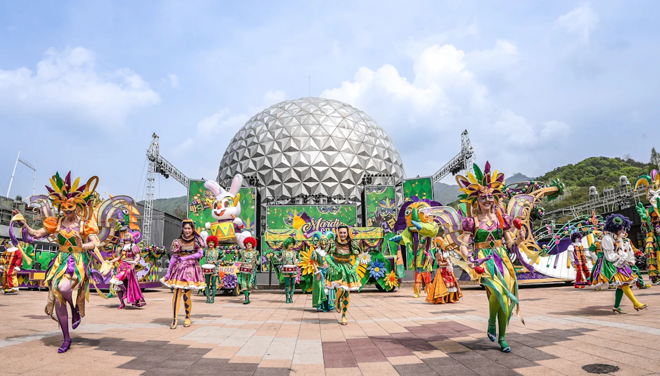 Performers in colorful costumes dancing in front of a geodesic dome and festive decorations under a blue sky.