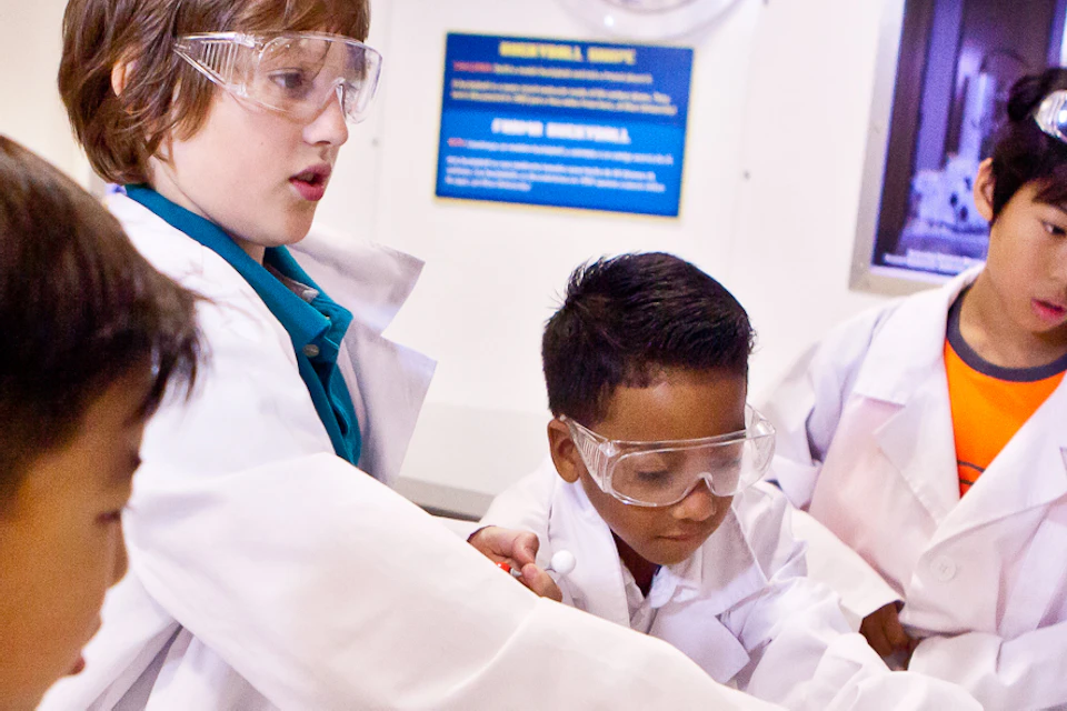 Children in lab coats and safety goggles work on a science experiment with molecular models and colored pebbles in a lab setting.