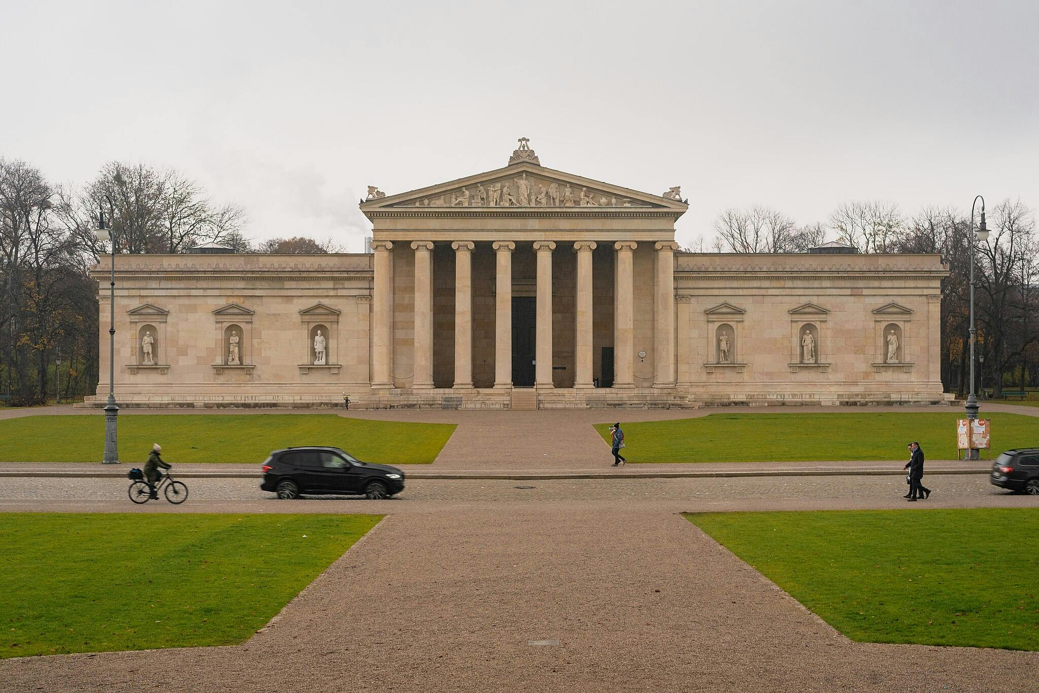 Edifici neoclàssic amb columnes i estàtues, gespa verda, gent caminant, un ciclista i cotxes en un dia ennuvolat.
