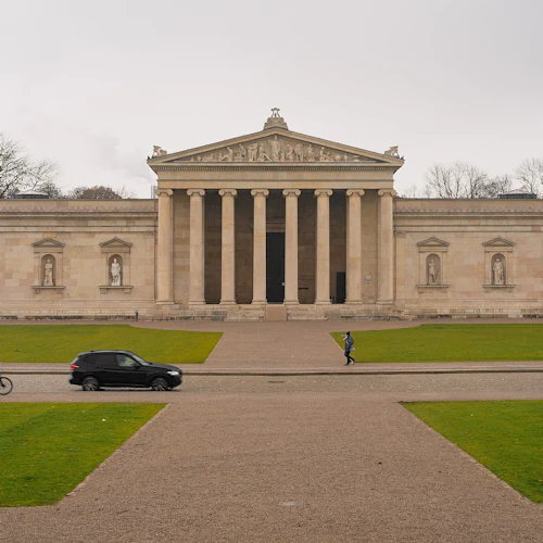 Neoklassizistisches Gebäude mit Säulen und Statuen, grüner Rasen, Spaziergänger, ein Radfahrer und Autos an einem bewölkten Tag.
