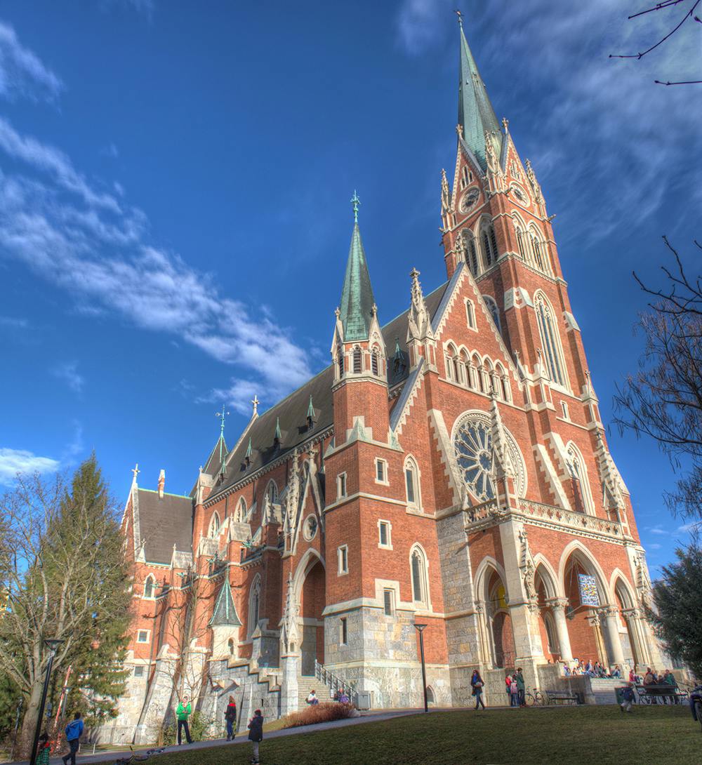 Una gran catedral d'estil gòtic de maó vermell amb torres altes i finestres arquejades sota un cel blau, amb gent passejant a prop.