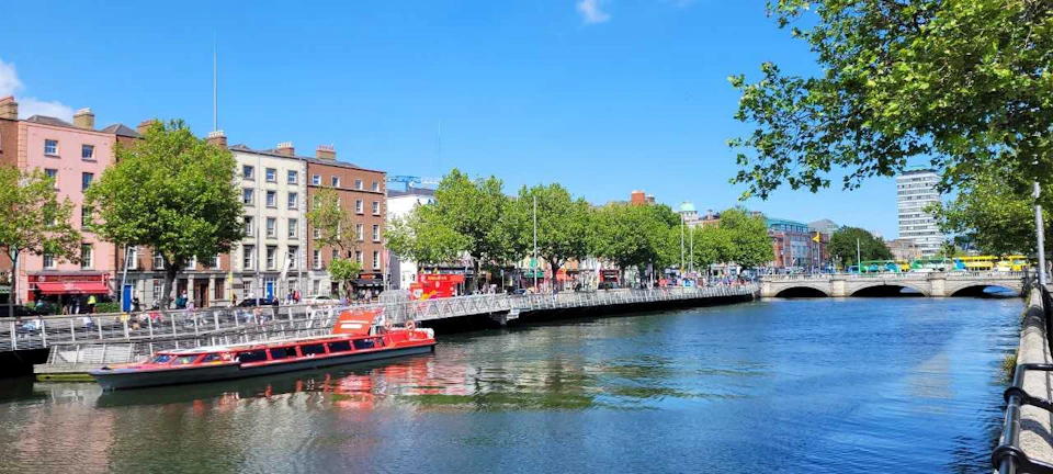 A riverside scene with a docked red boat, a bridge, trees, buildings, and a clear blue sky.