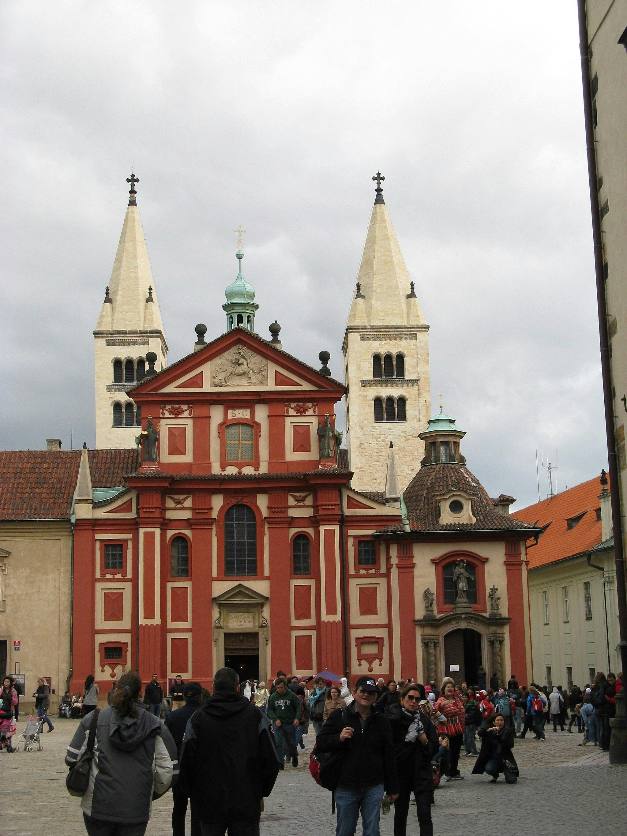 Le persone camminano davanti a un edificio storico rosso e beige con due alte torri in una giornata nuvolosa.