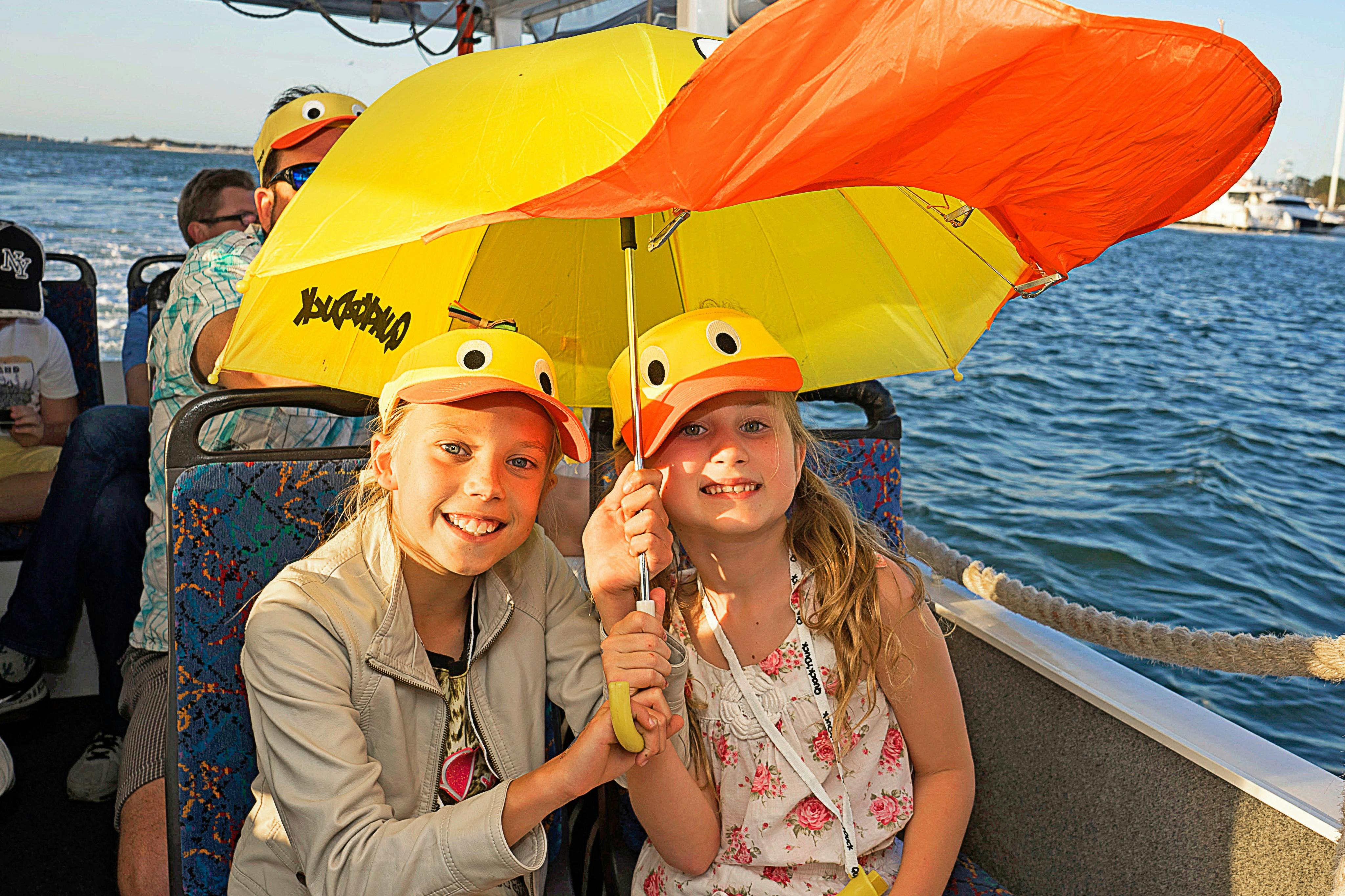 Dvě usmívající se děti se žlutými kachními klobouky sdílejí na lodi s ostatními cestujícími v pozadí stejný deštník s kachním motivem.