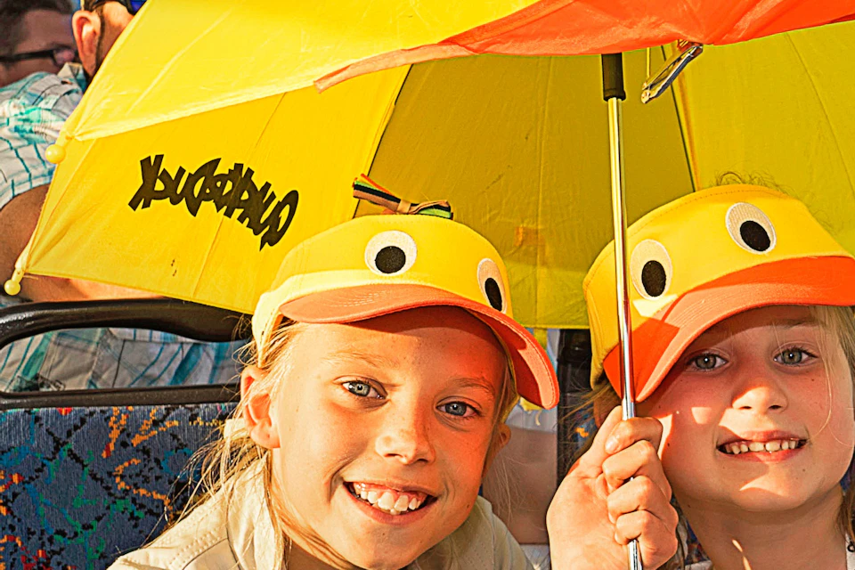 Two smiling children wearing yellow duck hats share a matching duck-themed umbrella on a boat with other passengers in the background.