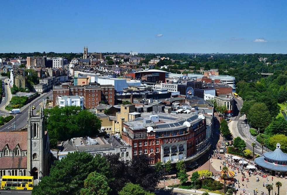 Aerial view of a town with a mix of historic and modern buildings, trees, and a church in the background under a clear blue sky.