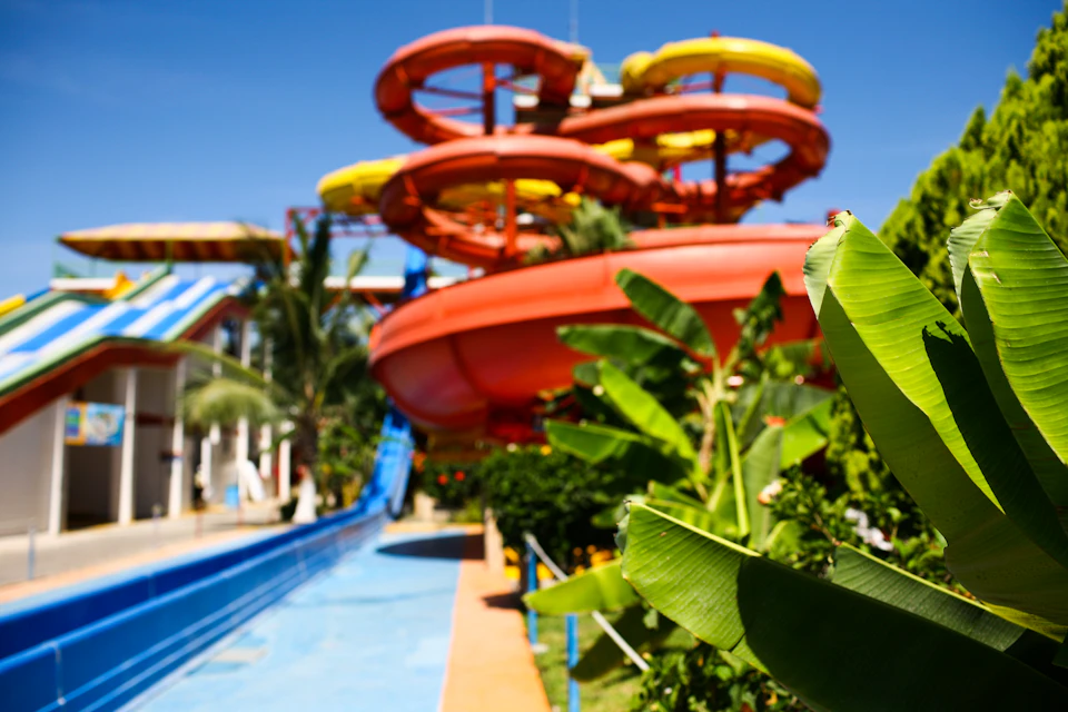 Brightly colored water slides with red, orange, and yellow tubes, surrounded by tropical plants and a clear blue sky.