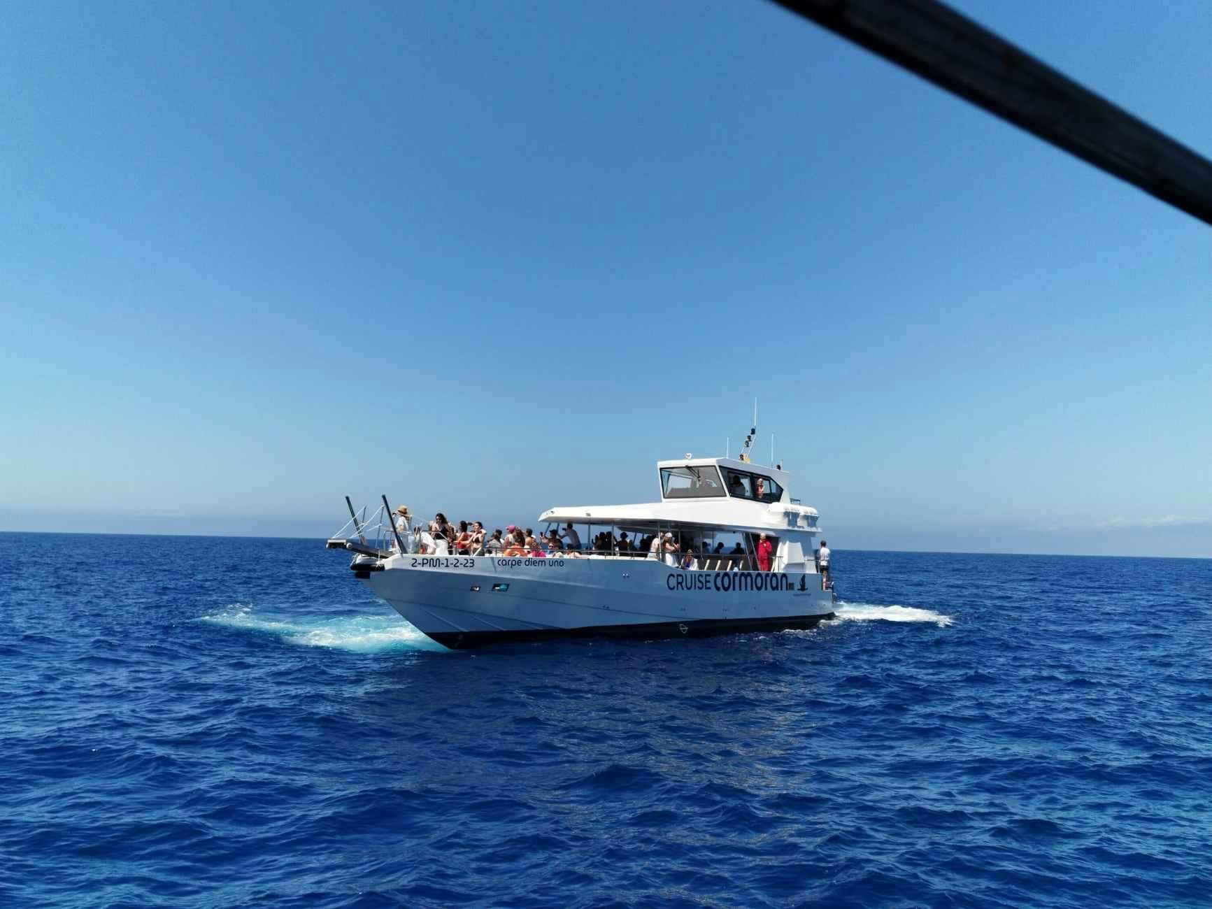 Una barca da crociera bianca che trasporta passeggeri naviga in un mare blu e calmo sotto un cielo limpido.