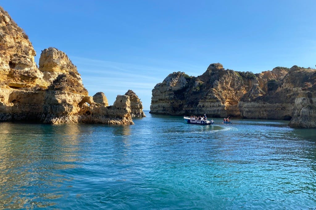 A small boat and a kayak with people navigate calm blue waters surrounded by rugged, sunlit cliffs under a clear sky.