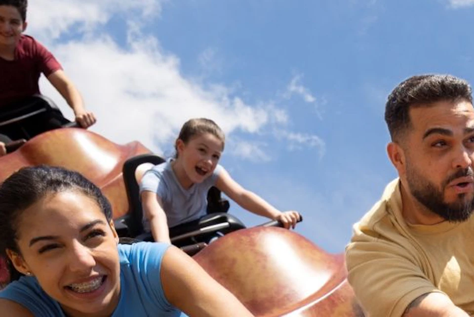People riding a rollercoaster with a vibrant, orange design, appearing excited and animated under a sunny sky.
