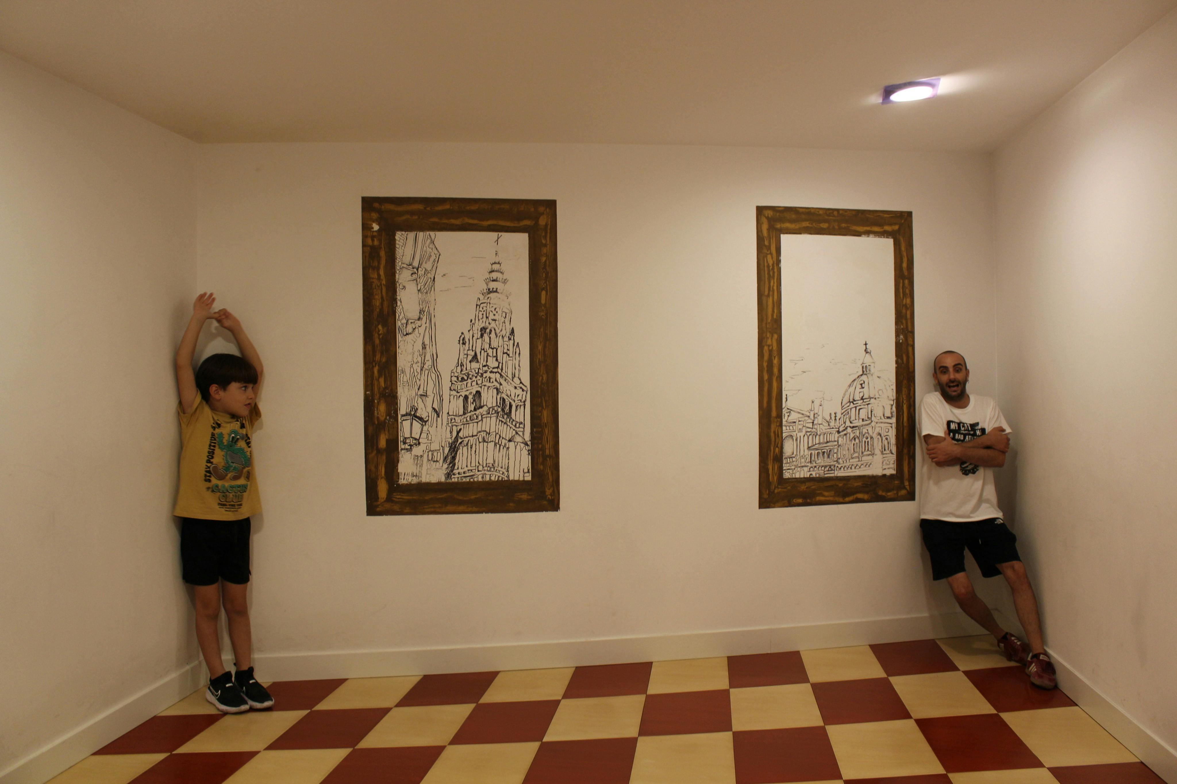 Un home amb roba esportiva es recolza contra una paret blanca amb esbossos arquitectònics emmarcats, de peu sobre un terra a quadres vermell i beix.