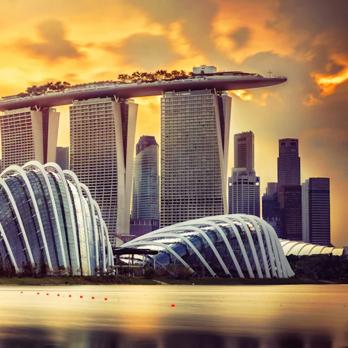Stadtbild mit futuristischen Gebäuden, darunter Marina Bay Sands und Supertree Grove, bei Sonnenuntergang mit Wasser im Vordergrund.