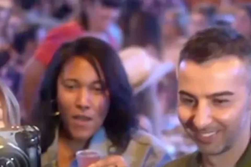 People raising glasses for a toast in a crowded, lively setting. One person is using a film camera to capture the moment.
