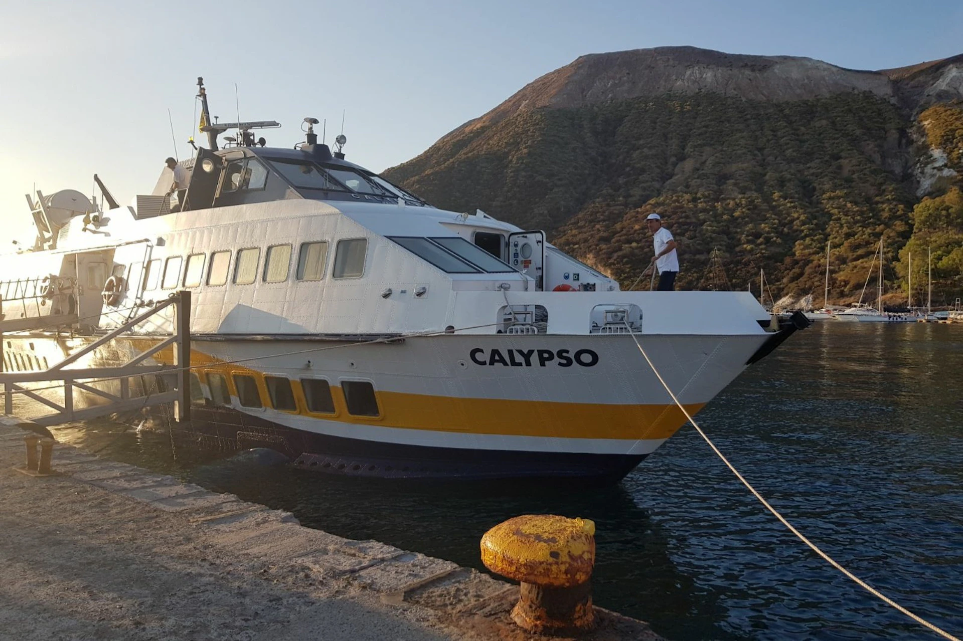 A boat named "CALYPSO" docked at a pier with a mountainous backdrop. A person is standing on the boat's deck.