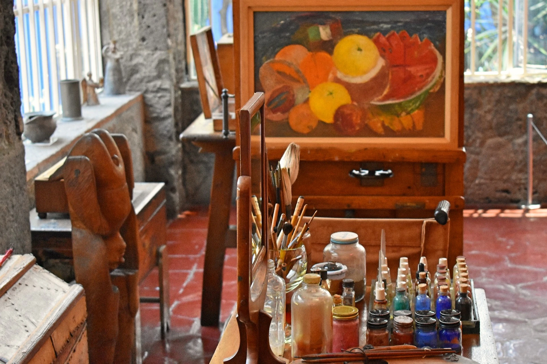 An artist's studio with paint bottles, brushes, and a palette. A colorful fruit painting is displayed on an easel in the background.