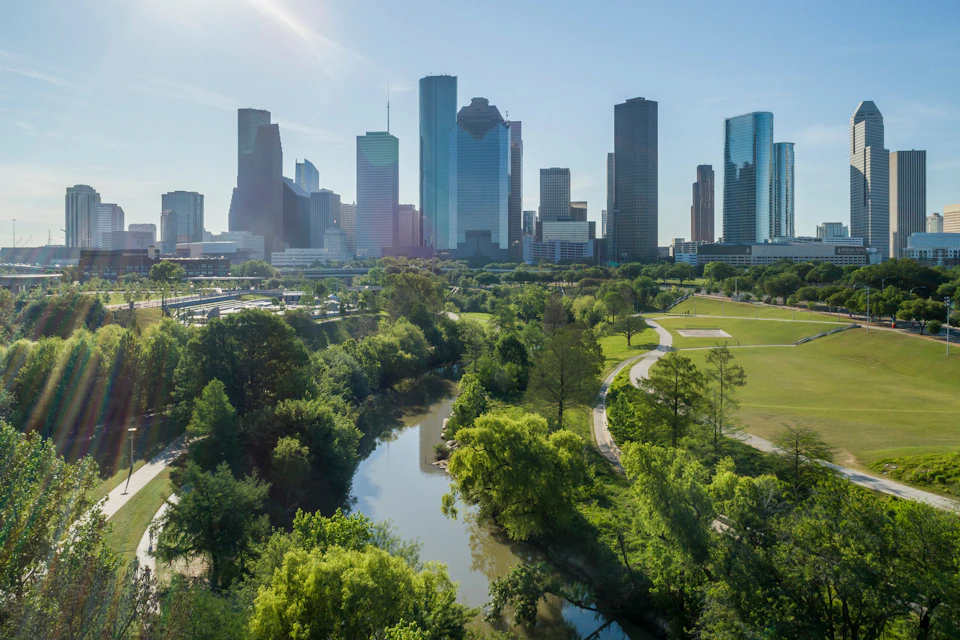 A cityscape of tall skyscrapers with a winding river and lush green park area in the foreground under a clear blue sky.