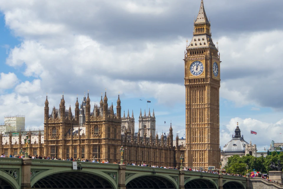 The Palace of Westminster and Big Ben with a crowded bridge in the foreground under a partly cloudy sky.