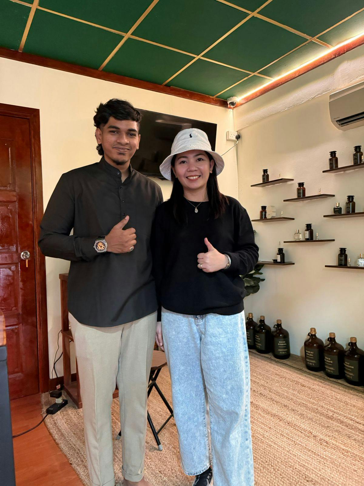 Two people pose indoors near shelves with bottles and a wooden door, one giving a thumbs-up gesture.