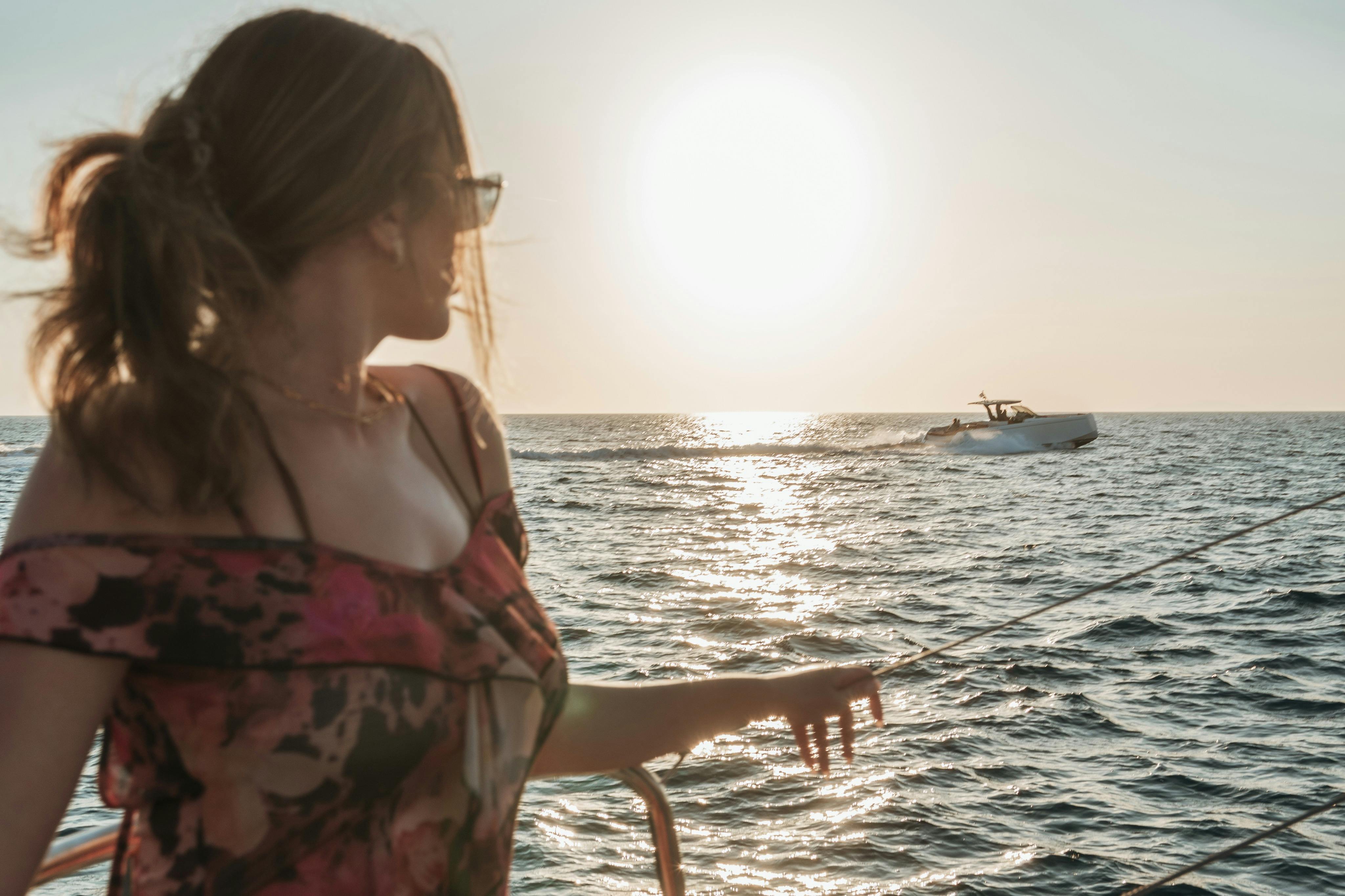A person with sunglasses and a floral dress looks towards the ocean at sunset, with sunlight reflecting on the water.