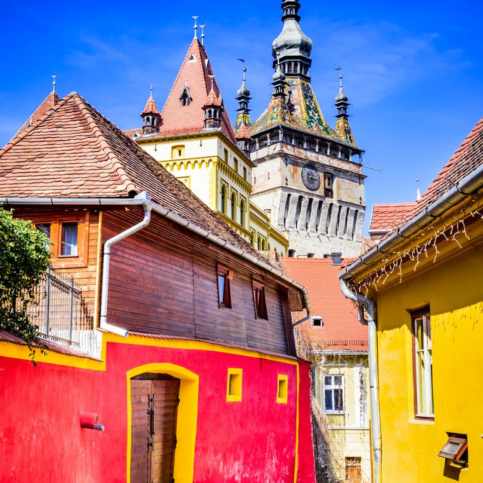 Colorful buildings under a blue sky, with a tall clock tower featuring spires and turrets in the background.