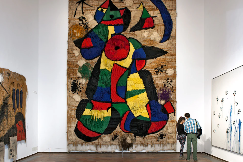 Two people observe a large, colorful abstract tapestry in an art gallery with white walls and wooden floor.