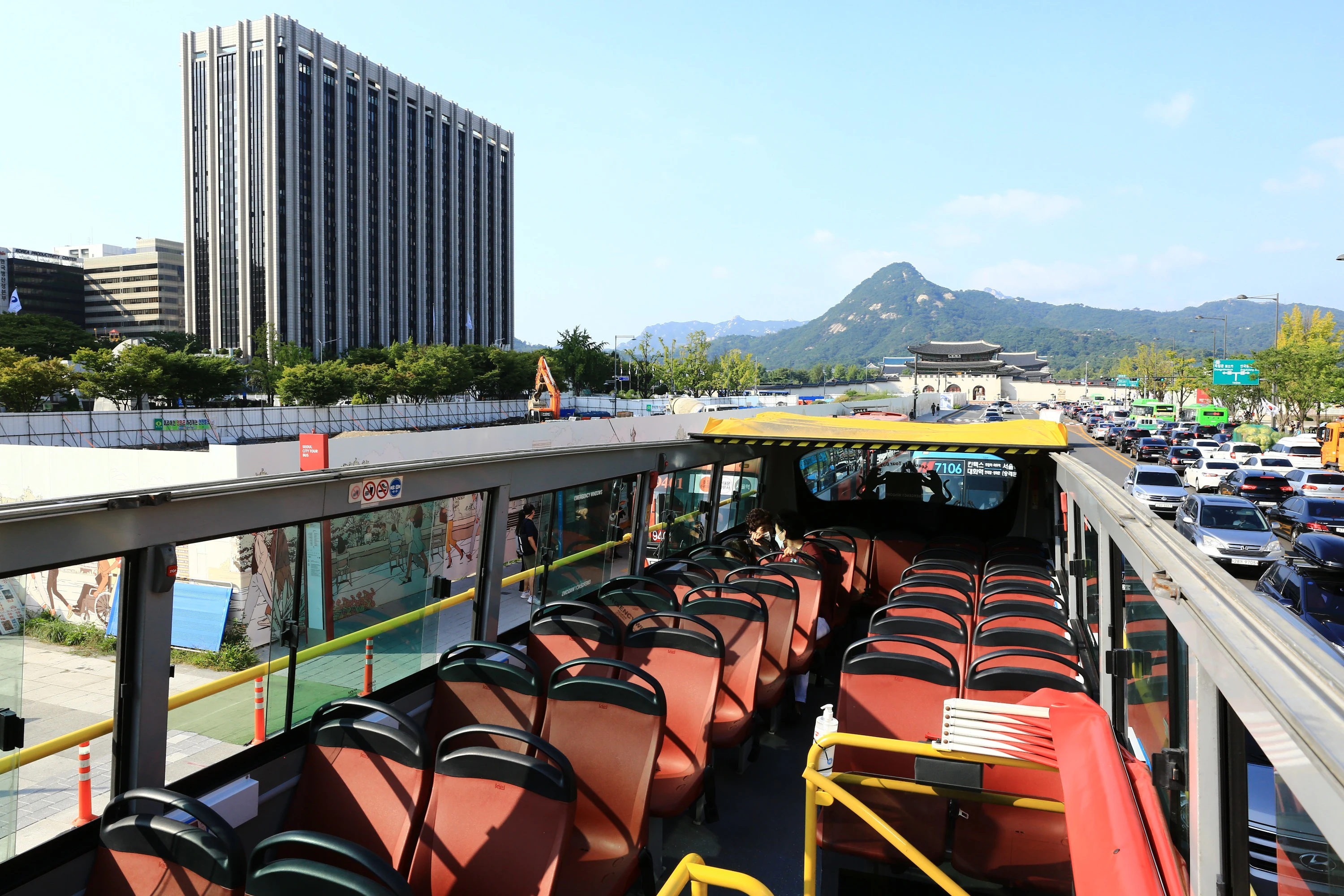 Open-top double-decker bus with empty red seats, a city building, and a distant mountain and gate under a clear blue sky.