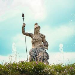 Stenstaty av en bepansrad figur med ett spjut omgivet av fontäner, blommor och grönska under en delvis molnig himmel.