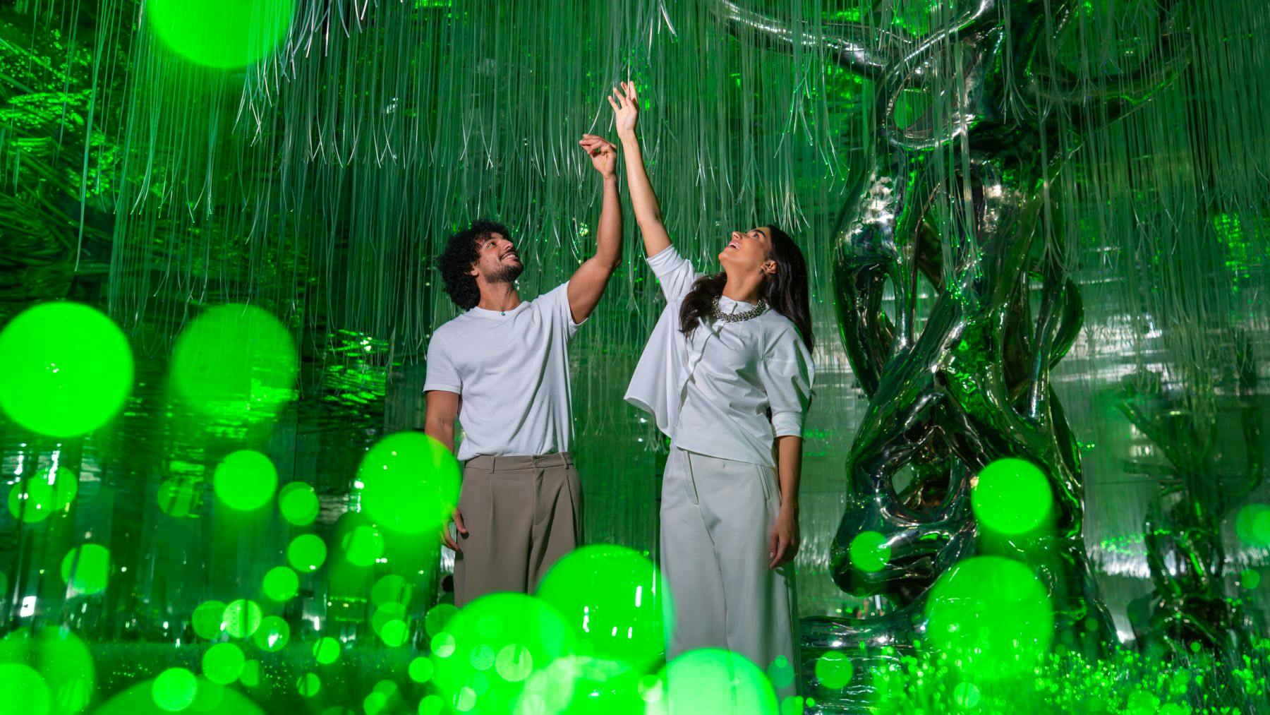 흰색 의상을 입은 두 사람이 녹색 조명, 반사 표면, 행잉 스트링이 있는 방에서 위쪽으로 손을 뻗고 있습니다.
