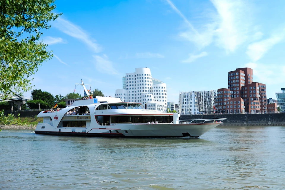 A modern boat is cruising along a river with contemporary buildings in the background under a clear blue sky.