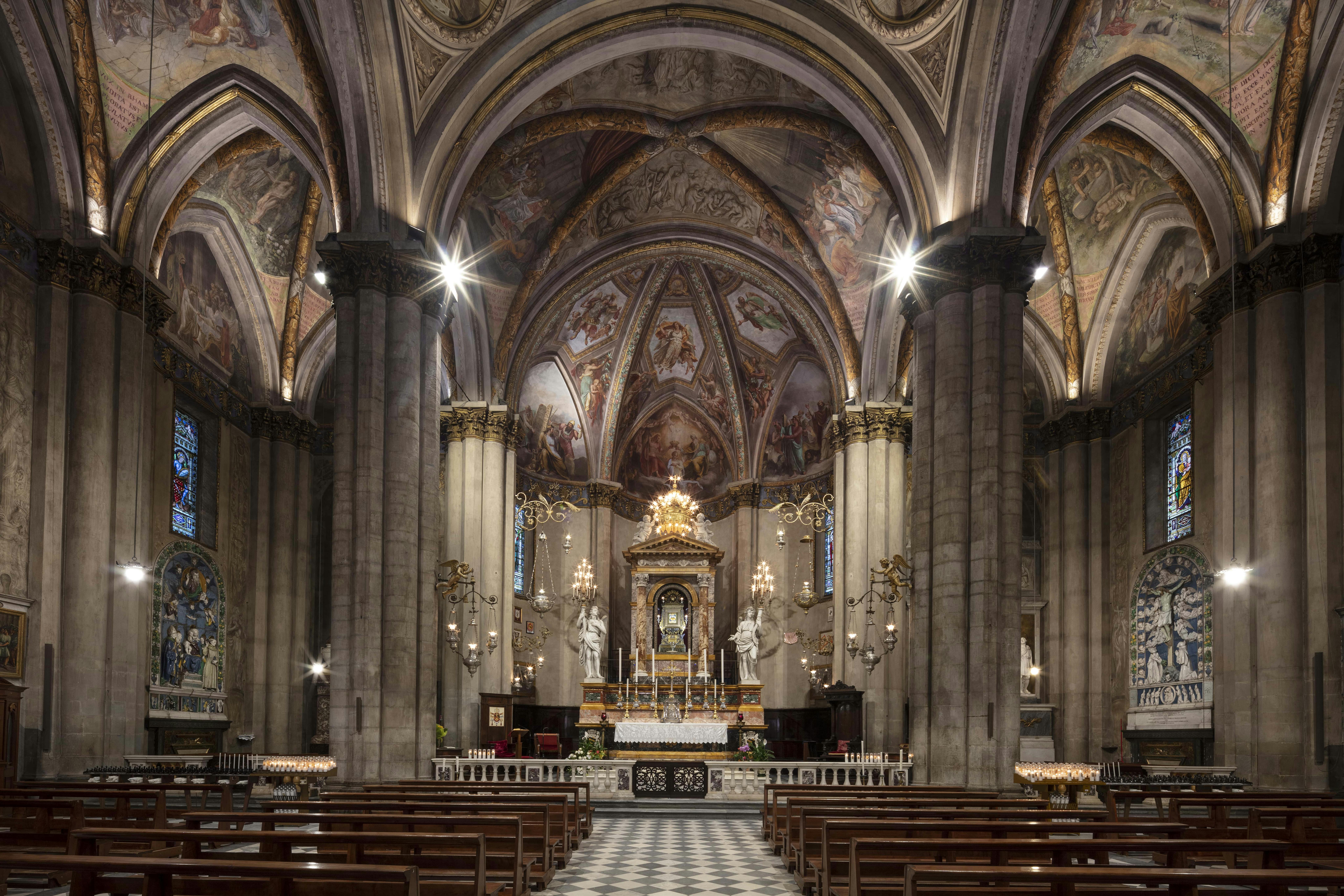 Un interno di cattedrale con archi decorati, murales, colonne, banchi e un altare decorato con statue e candele.