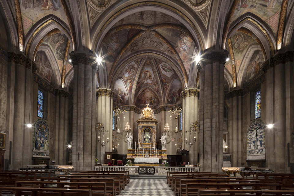 A cathedral interior with ornate arches, murals, columns, pews, and a decorated altar with statues and candles.