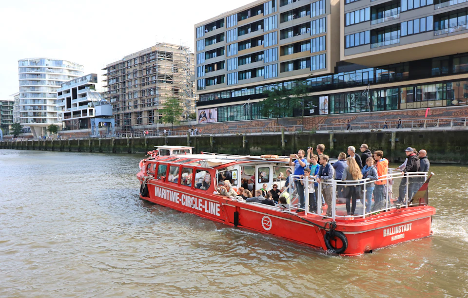 A red tour boat labeled "Maritime Circle Line" travels through a river, with passengers on board, adjacent to modern waterfront buildings.