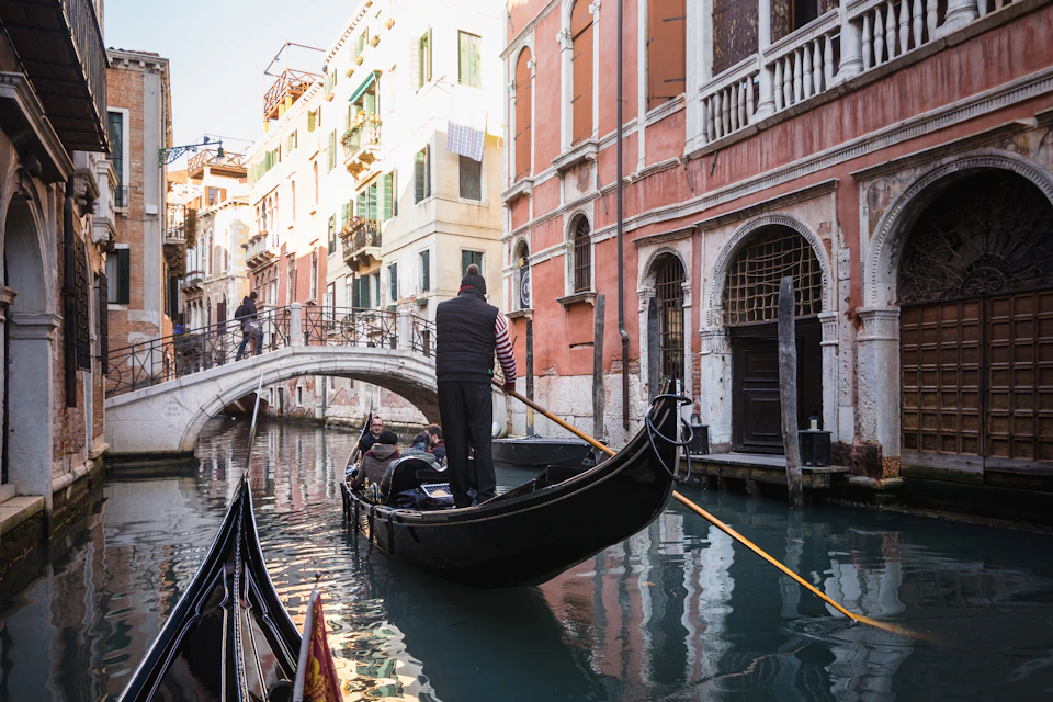 A gondolier navigates a gondola with passengers on a canal, with a stone bridge and historic buildings lining the waterway.