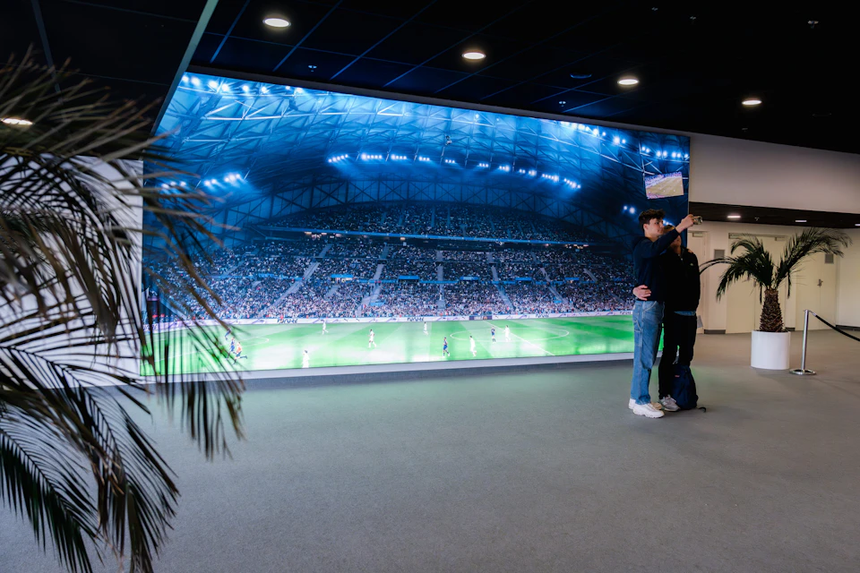 A large screen displays a lit-up stadium during a soccer match. Two people stand nearby in an indoor setting.