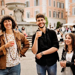 Three people smiling and holding drinks in a lively outdoor plaza with a fountain and historic buildings in the background.